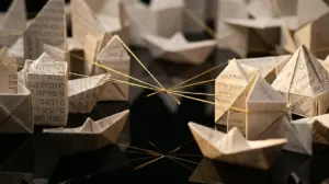 אוריגמי של סירות ומבנים מנייר עם טקסט רב-לשוני, המחוברים ברשת של חוטי זהב על משטח השתקפות כהה.